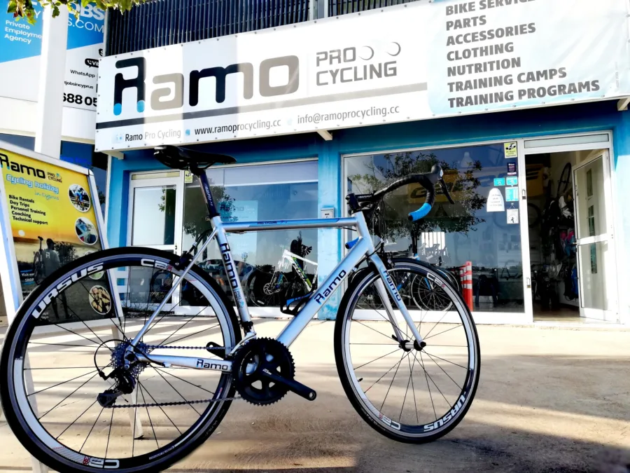 A silver Ramo road bike on its stand outside the shop, under the Ramo Pro Cycling banner listing bike service, parts, clothing and training camps
