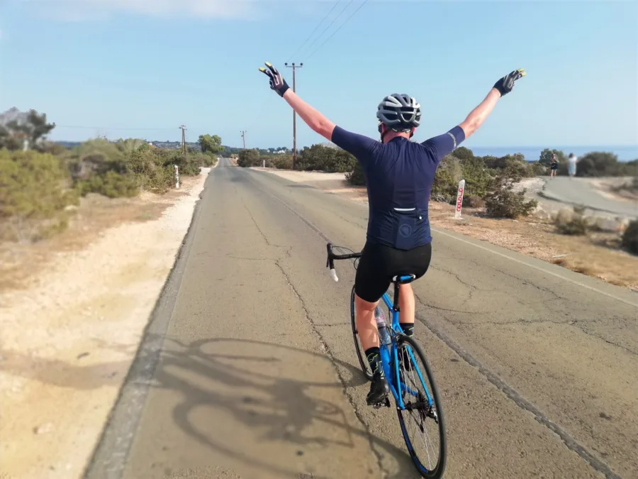 A rider from behind with both arms thrown wide on an empty coastal road, the sea on the horizon