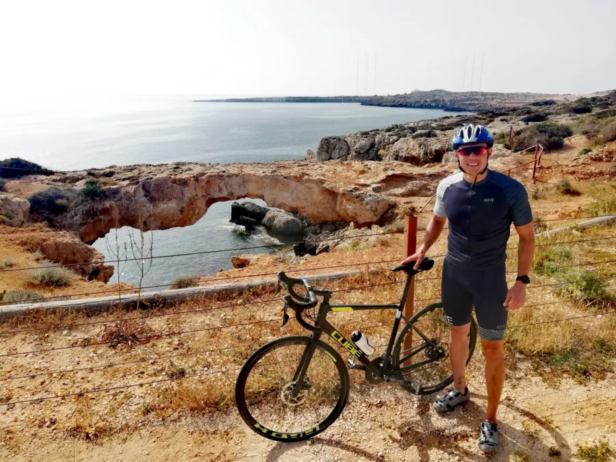 A rider standing with a road bike above a natural rock arch on the coast