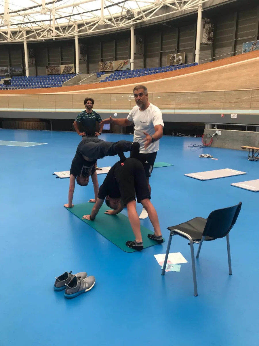 A coach guiding two people through a stretch on mats on the infield of an indoor velodrome