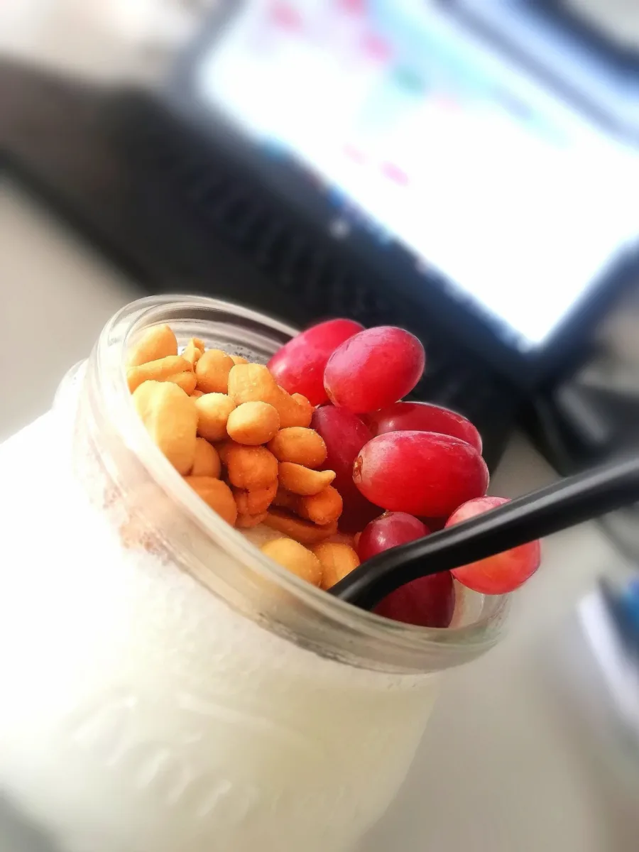 A jar of yoghurt topped with peanuts and red grapes, standing on a desk in front of a screen