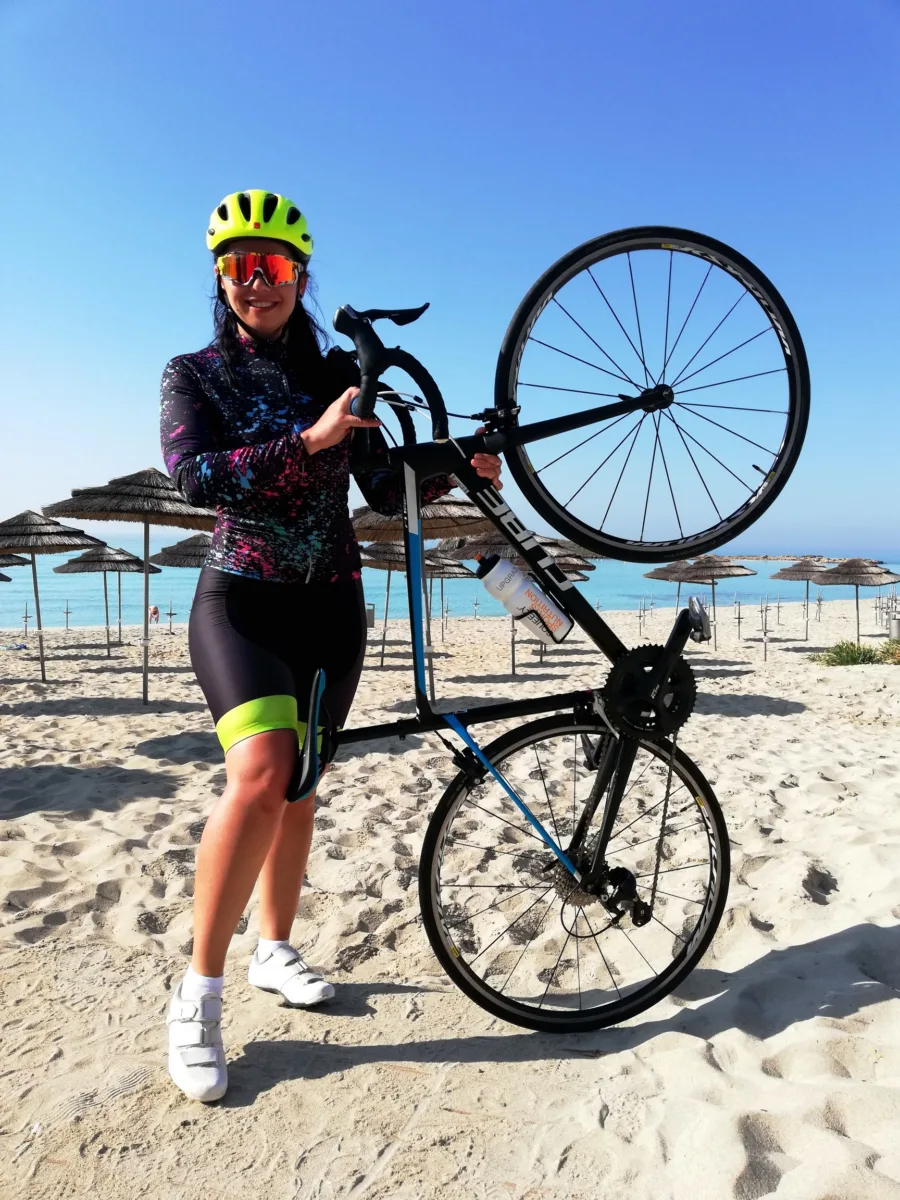 A rider holding a road bike upright on the sand in front of a line of straw parasols