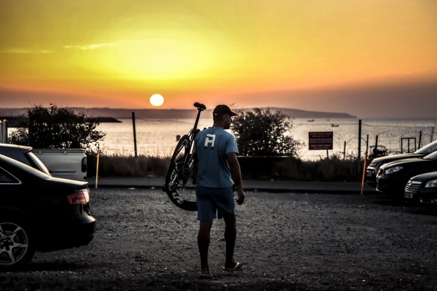 A rider carrying a bike over one shoulder across a gravel car park as the sun sets over the sea