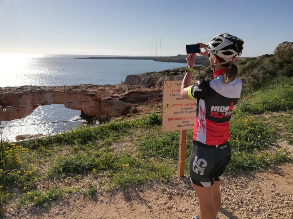 A cyclist in a helmet photographing a natural rock arch and the open sea from the clifftop at Cape Greco