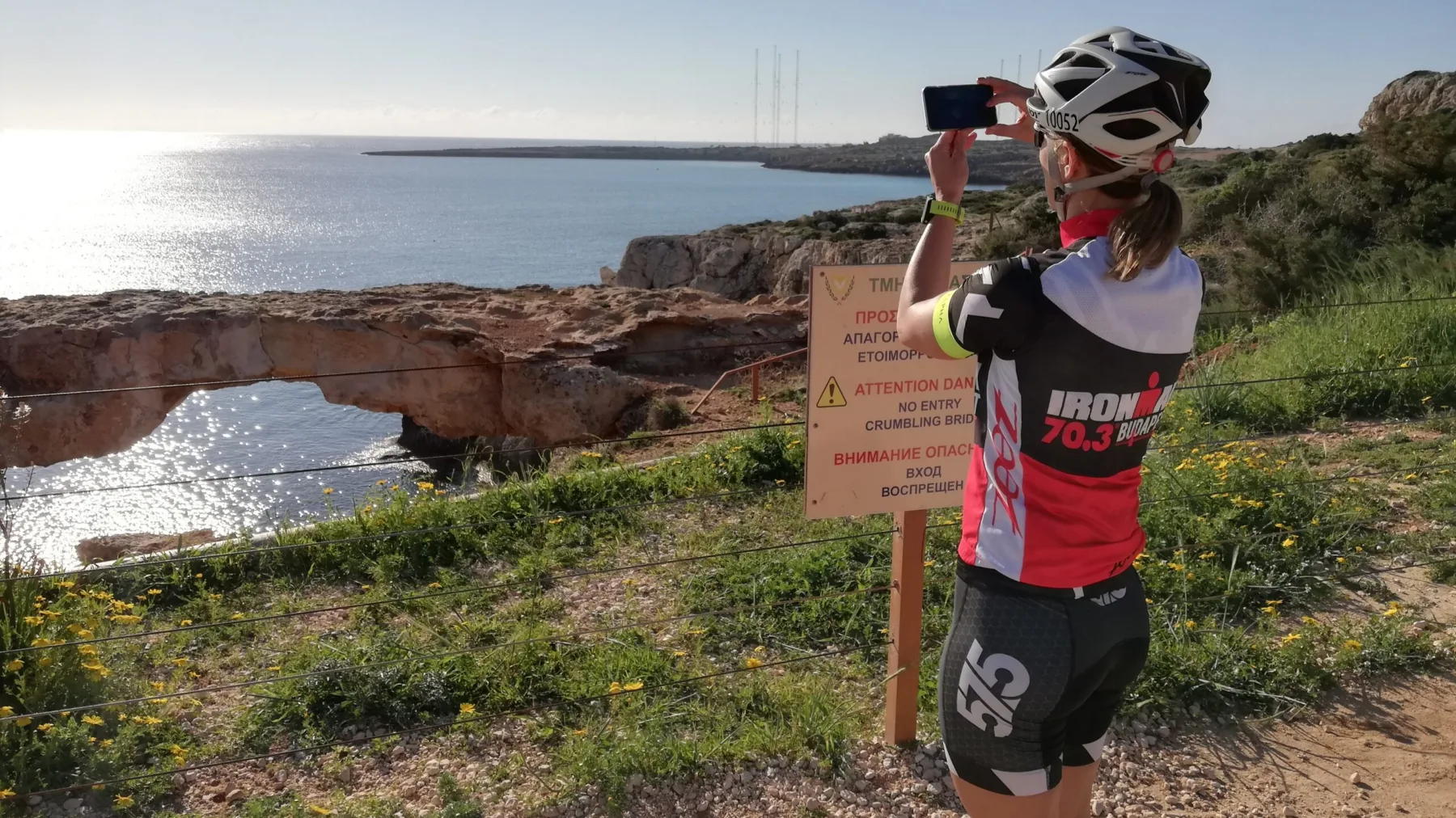 A cyclist in a helmet photographing a natural rock arch and the open sea from the clifftop