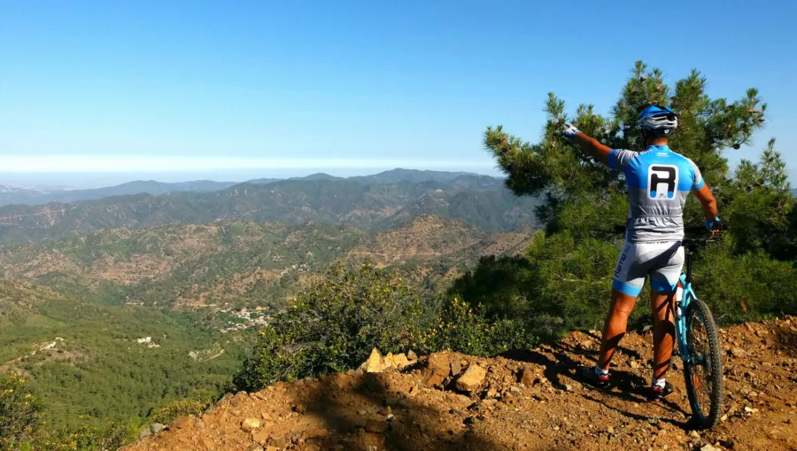 A mountain biker on a rocky spur pointing across a wide forested valley