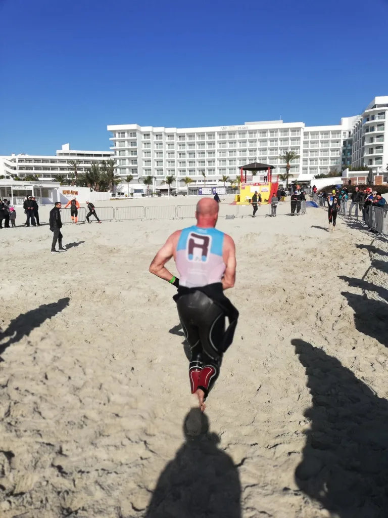 A triathlete in a Ramo top, wetsuit peeled to the waist, running up the beach towards a row of seafront hotels
