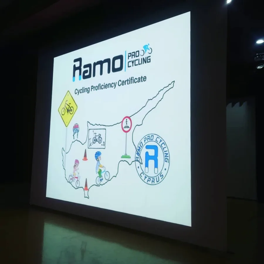 A projected slide reading Cycling Proficiency Certificate over a map of Cyprus drawn with road signs and young riders