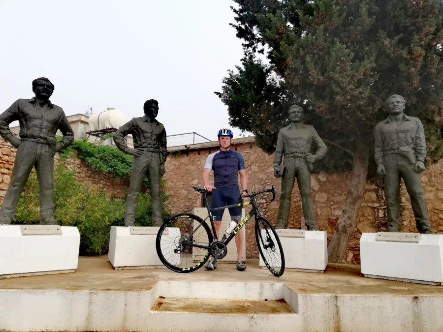 A rider standing with a road bike between four bronze figures on plinths at a village memorial