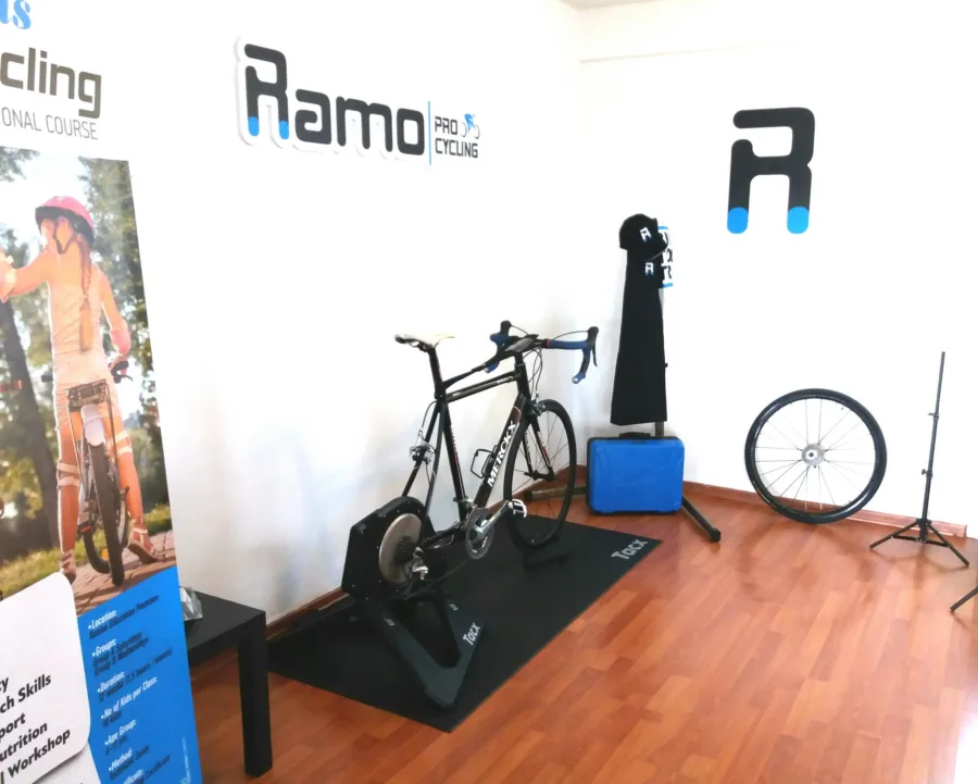 A road bike on a turbo trainer in the studio, a spare wheel and a roll-up banner for the kids cycling course beside it