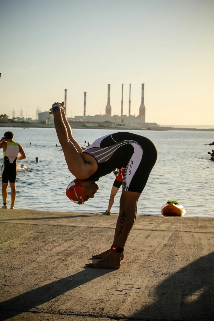 A triathlete folded double stretching on a slipway at dawn, chimneys across the bay behind