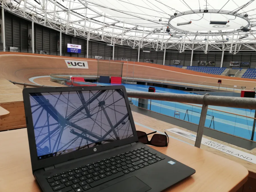 A laptop open on a desk overlooking an indoor wooden velodrome, the UCI logo on the boards