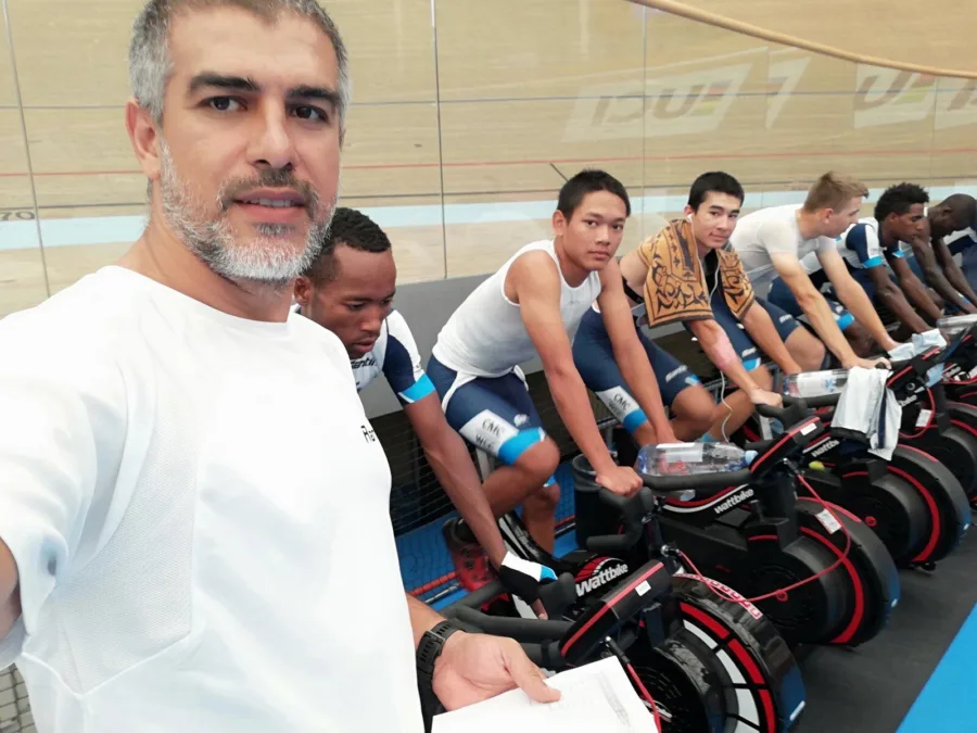 A line of riders on turbo trainers on a velodrome infield beneath a UCI banner