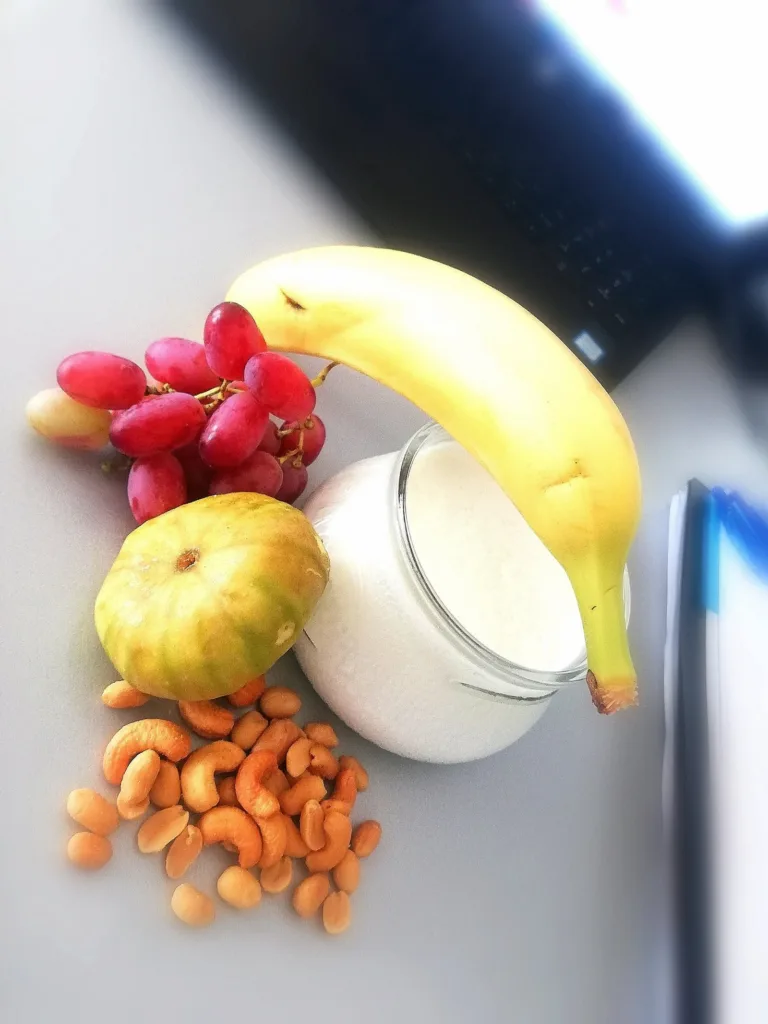 Banana, red grapes, a fresh fig, cashews and a tub of yoghurt laid out on a white surface