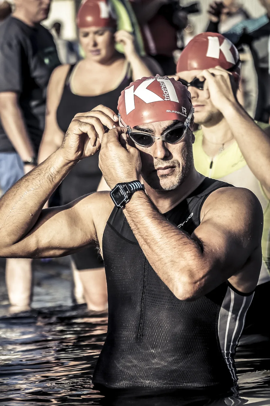 Triathletes in red swim caps pulling on their goggles in the water before a swim start