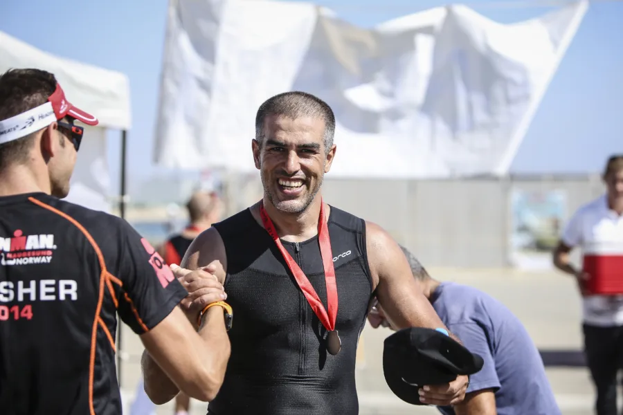 Two men clasping hands at a race finish, one in a tri suit wearing a medal, an Ironman finisher banner behind