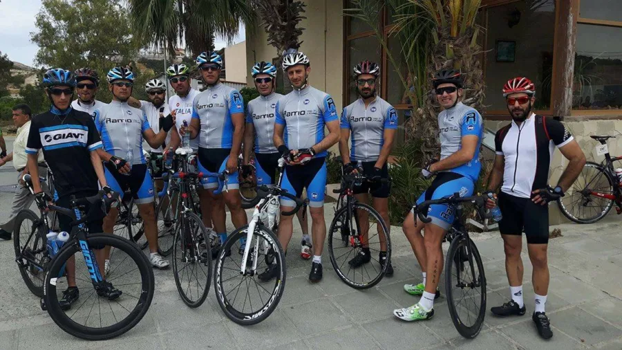 Nine riders in blue Ramo team kit standing in a line with their road bikes in front of a low building