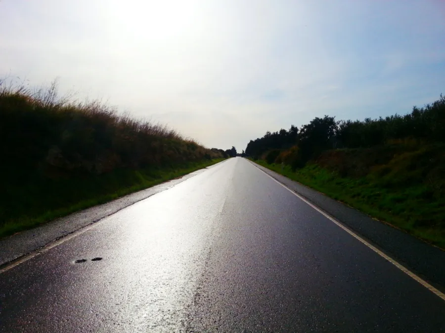 A wet empty road running between grass banks under a bright hazy sky