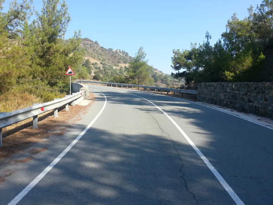 A mountain road curving uphill between pines behind a crash barrier
