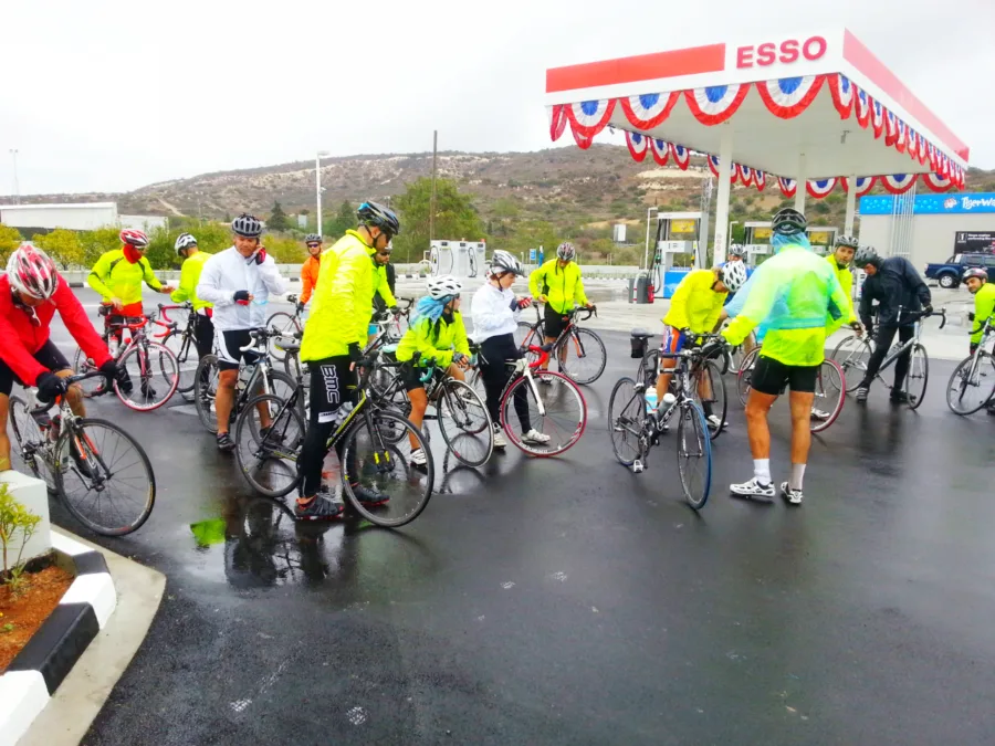 A big group of riders and bikes gathered under the canopy of an Esso station in the rain