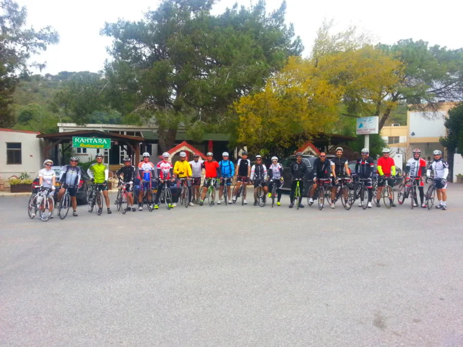 A large group of riders and bikes gathered on gravel beside a roadside kiosk