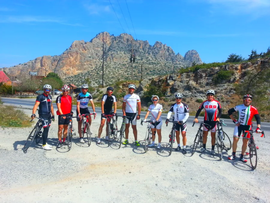 A group of riders with their bikes on gravel below a jagged rock peak