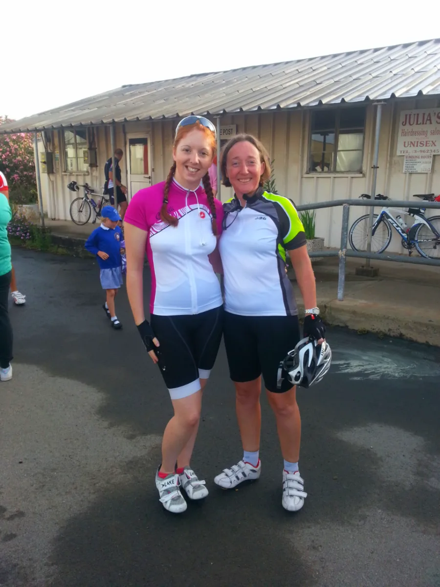 Two riders in cycling kit standing together outside a building with bikes behind