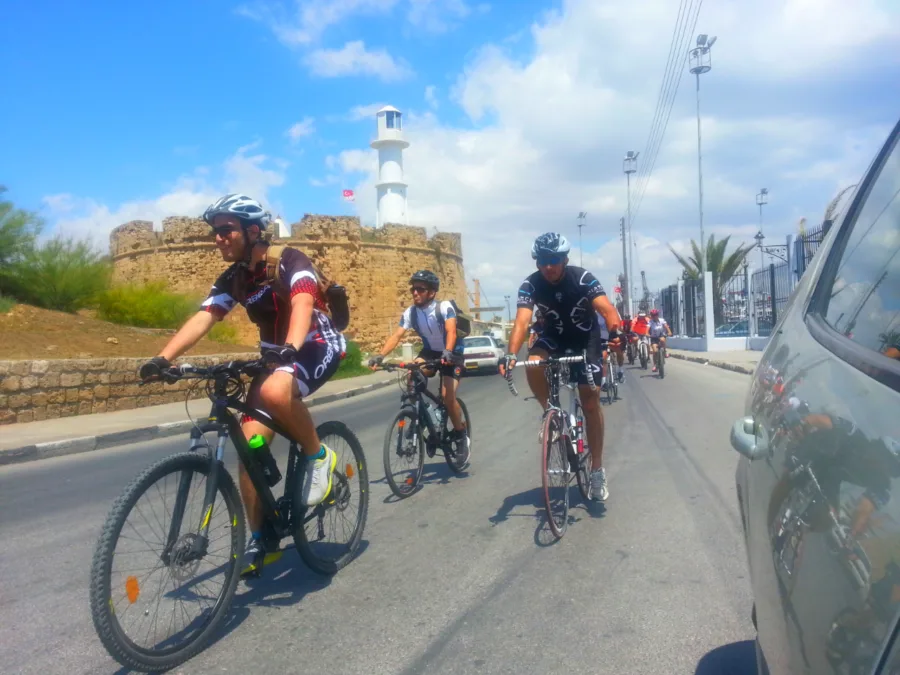 Riders on a seafront road passing a stone fort and a lighthouse