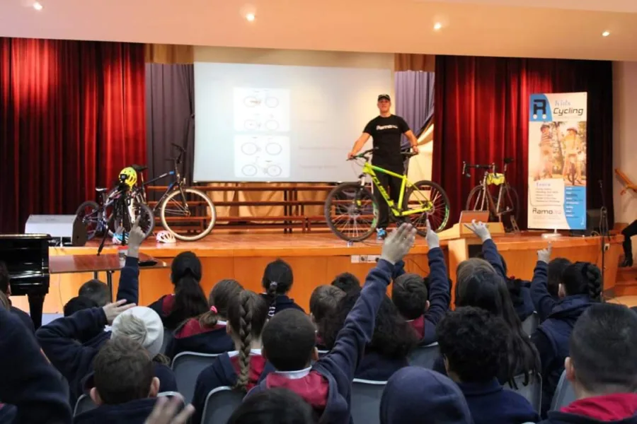 Ramo on a school stage beside a mountain bike and a road bike, presenting to a hall of schoolchildren with their hands raised