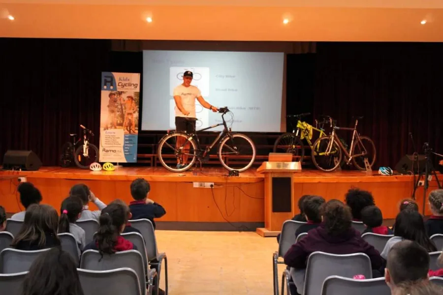 A school stage lined with bicycles and a presentation screen, children seated in front