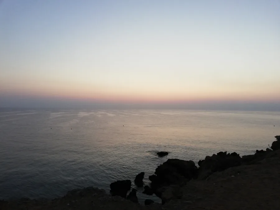 A pink and gold sky over a flat calm sea and dark rocks