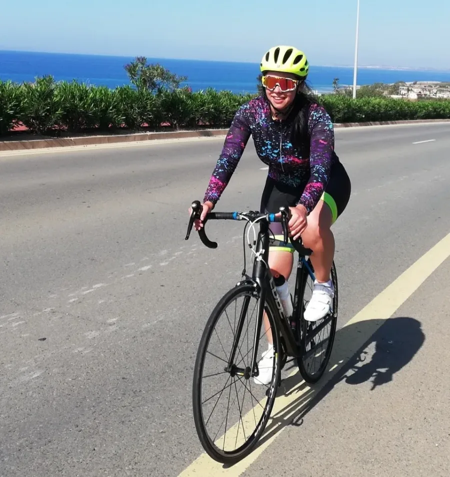 A rider on a road bike smiling on a wide coast road, the sea and a headland behind